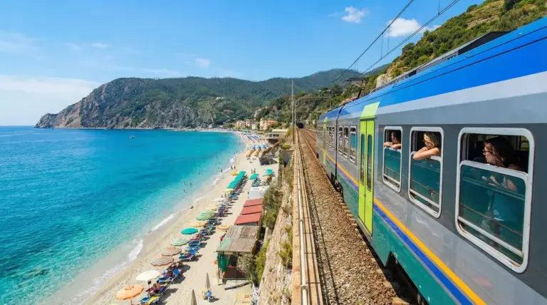 Treno che costeggia una spiaggia con mare azzurro e ombrelloni colorati